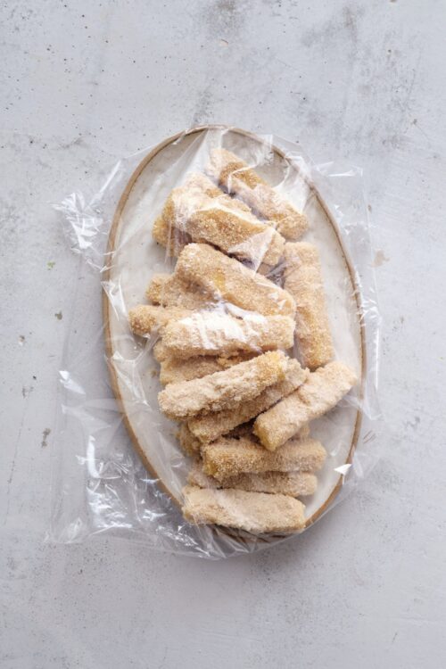 Coconut breaded chicken tenders on a white platter with plastic wrap, ready for baking or frying.