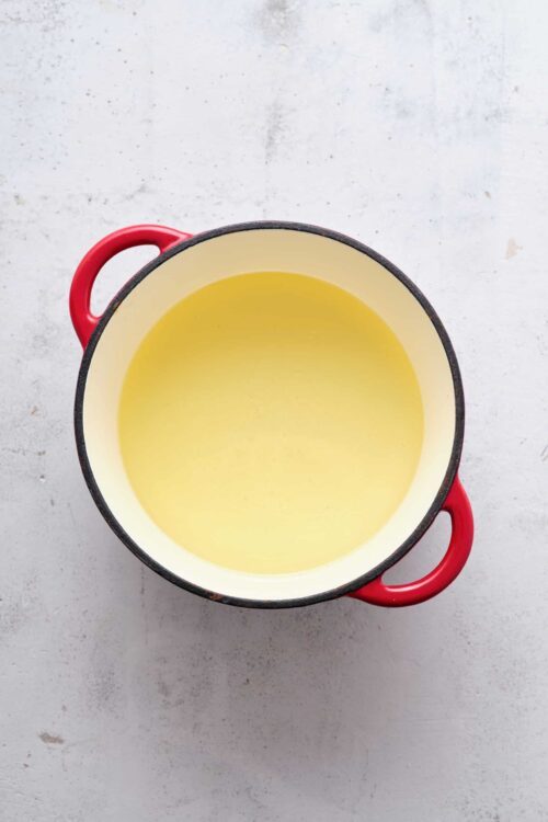 Cream-colored pot with red handles containing melted butter or oil, on a light textured surface.
