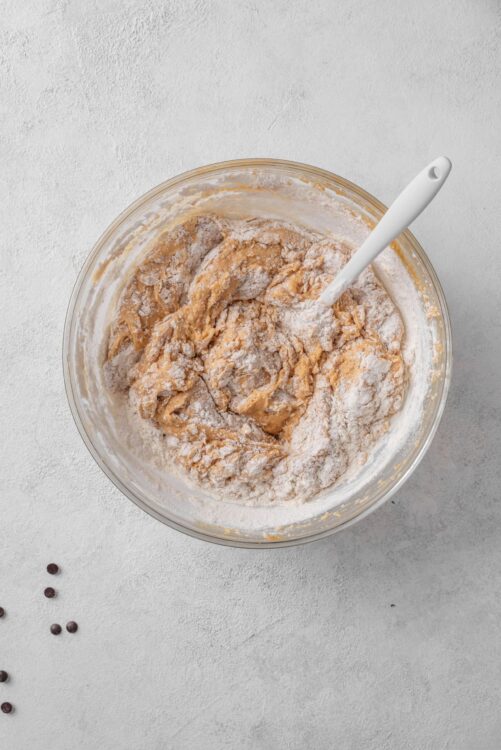 Creamy cookie dough mixture in glass bowl with spatula, ready for baking, on light textured surface.