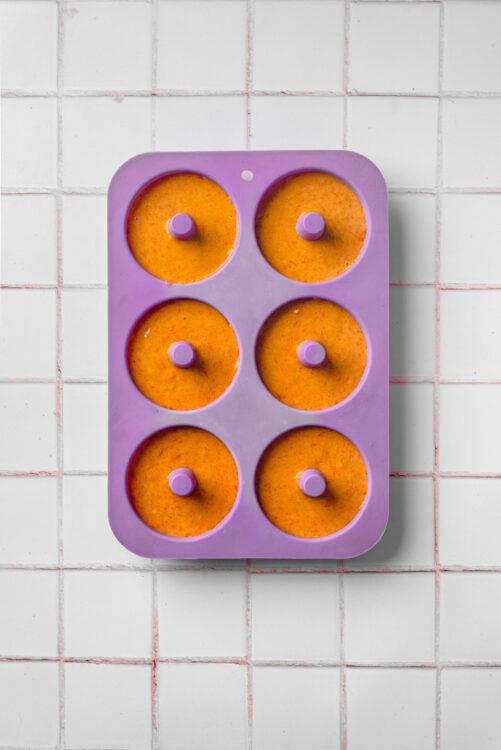Bright orange frozen fruit puree in purple silicone pop mold on white tiled background.