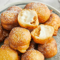 Golden fried cream-filled doughnuts dusted with powdered sugar, served on a decorative plate.