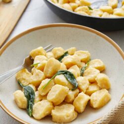 Creamy gnocchi with sage leaves served in a beige bowl on a light surface. Fresh gnocchi with melted butter and sage garnish.