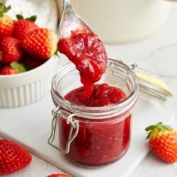 Fresh homemade strawberry jam in a glass jar with a spoonful being lifted out, with fresh strawberries around it.