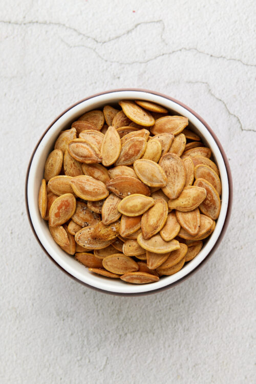 Roasted salted pumpkin seeds in a white bowl on textured surface.