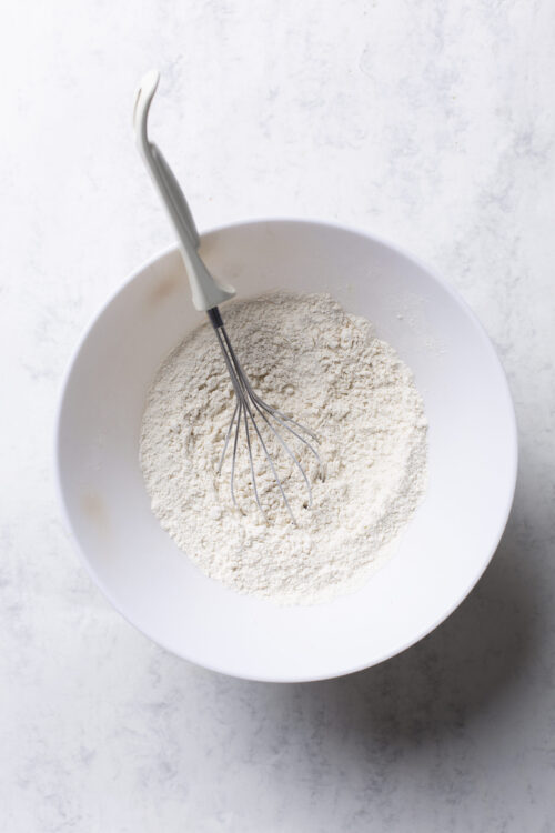 Flour being mixed in a white bowl with a whisk for baking recipes.