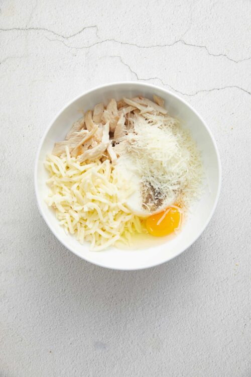 Shredded chicken, cheese, eggs, and seasonings in a white bowl for baked chicken casserole.