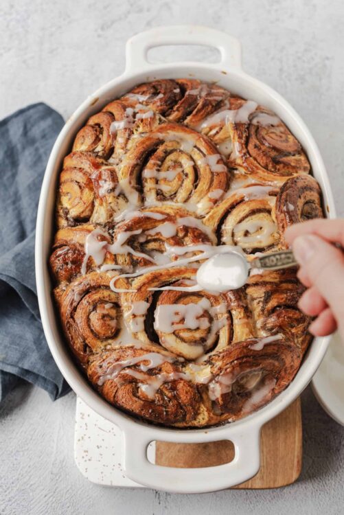 Cinnamon roll bake in a white baking dish drizzled with icing, served as a sweet breakfast treat.