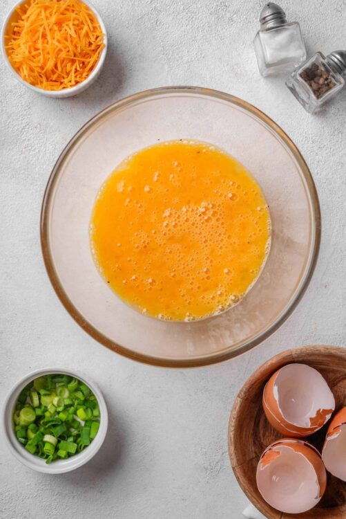 Egg mixture in glass bowl with chopped green onions, shredded cheese, and cracked eggs around it.