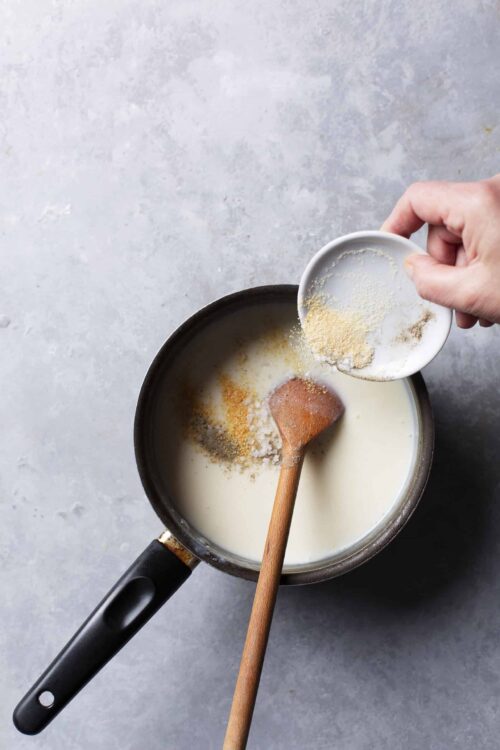 Creamy sauce being prepared with spices in a saucepan on a gray surface.