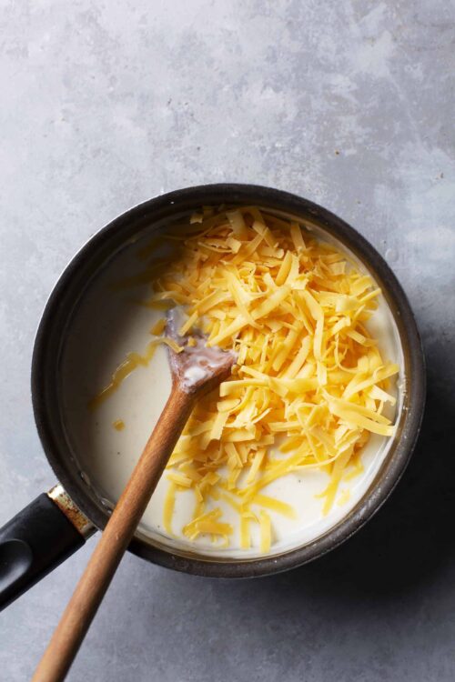 Creamy cheese sauce with shredded cheddar cheese melting in a saucepan.