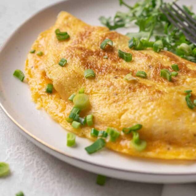Golden cheese omelette garnished with chopped green onions on a white plate with fresh greens in the background.