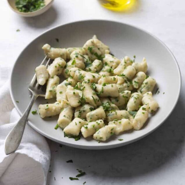 Creamy gnocchi with herbs on a white plate, ready to serve, with olive oil and herbs in the background.