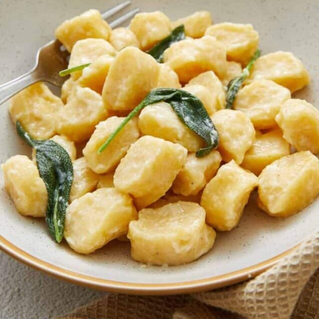 Creamy gnocchi with sage leaves served in a beige bowl on a light surface. Fresh gnocchi with melted butter and sage garnish.