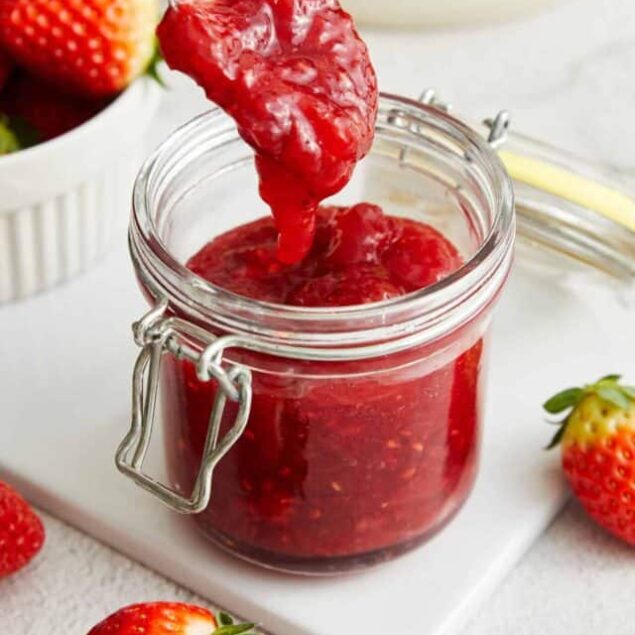 Fresh homemade strawberry jam in a glass jar with a spoonful being lifted out, with fresh strawberries around it.