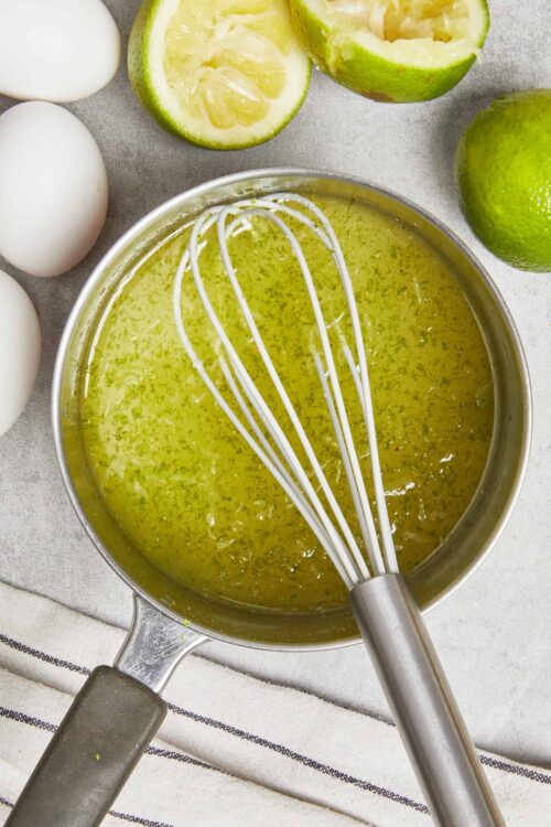 Lemon-lime juice in a saucepan with a whisk, surrounded by eggs and fresh limes on a kitchen countertop.