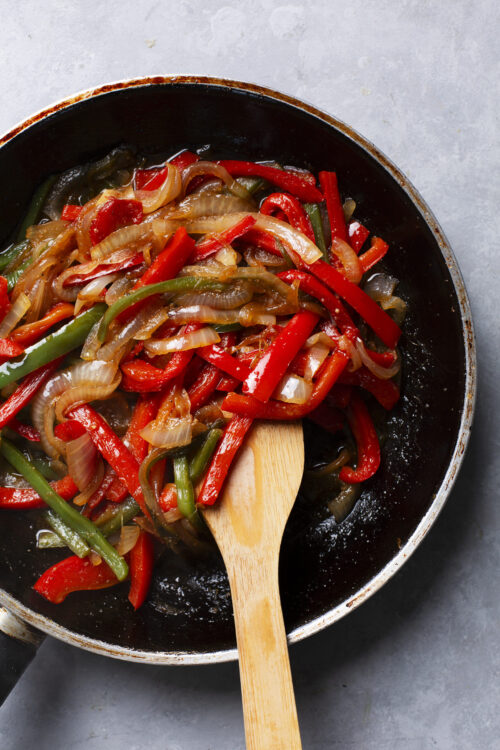 Sautéed colorful bell peppers and onions in a black skillet with a wooden spoon.
