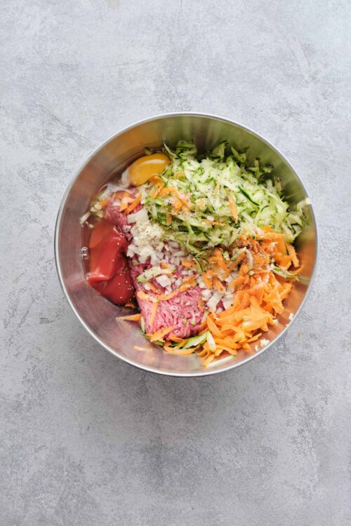 Chopped vegetables and raw ground beef in a stainless steel mixing bowl for meatloaf preparations.