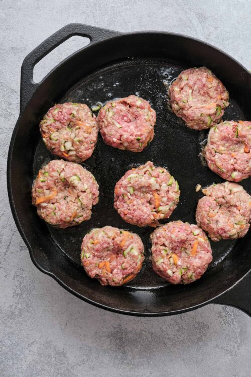 Seasoned meatballs cooking in a cast iron skillet with chopped vegetables.