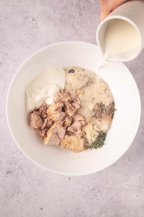 Cream of mushroom soup with tuna, sour cream, black pepper, onion, and herbs in a white bowl with milk being poured.