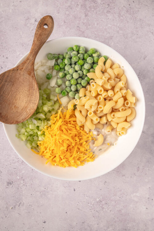 Colorful pasta salad ingredients in a white bowl with a wooden spoon, featuring peas, celery, cheese, and pasta.