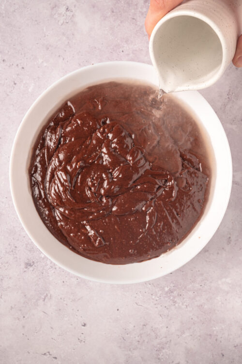 Rich chocolate brownie batter being mixed in a white bowl with cream poured over the mixture for a baking recipe.