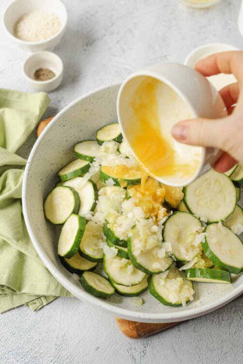 Sliced zucchini and chopped onions in a bowl with eggs being poured over.