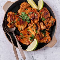 Golden grilled chicken thighs with lime wedges on a black skillet, topped with chopped herbs for a delicious meal.