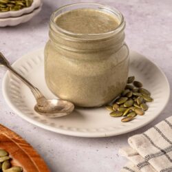 Creamy pumpkin seed butter in a glass jar on a white plate with scattered pumpkin seeds, gray background.