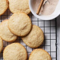 Buttery sugar cookies cooling on a wire rack with a bowl of sugar and cinnamon sticks nearby.