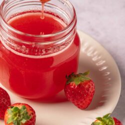 Fresh strawberry jam in a glass jar with whole strawberries on a white plate, perfect for baking and breakfast.