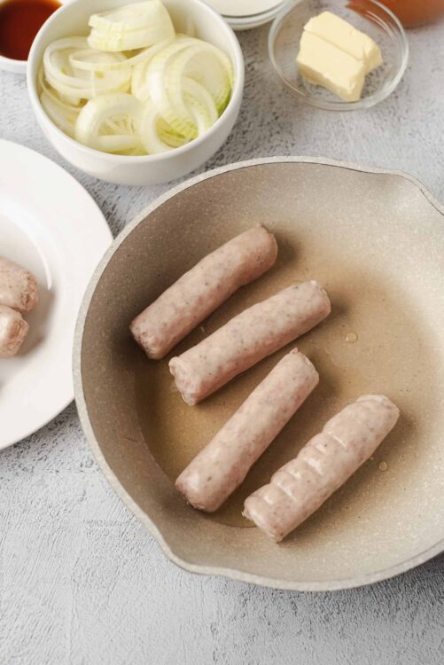 Raw sausages cooking in a skillet with sliced onions, butter, and seasonings for homemade sausage recipes.