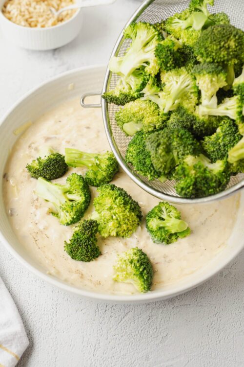 Fresh broccoli florets being added to a creamy cheese sauce in a white bowl.