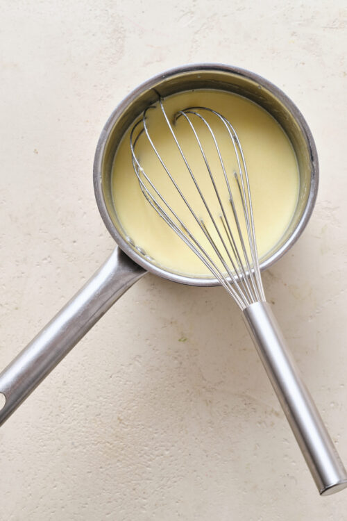 Creamy sauce in a stainless steel saucepan with a whisk on a textured beige surface.