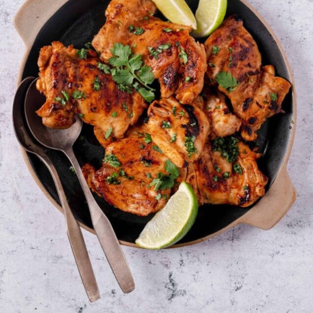 Juicy grilled chicken thighs garnished with cilantro and lime wedges in a black skillet.