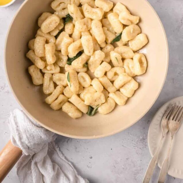 Creamy gnocchi in a white bowl garnished with sage leaves, served with lemon on the side.