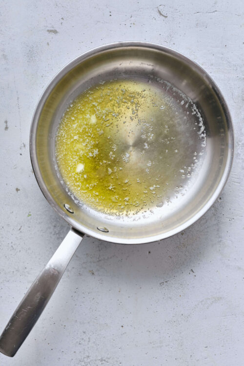Bright stainless steel saucepan with melted butter and garlic, on a white textured surface.