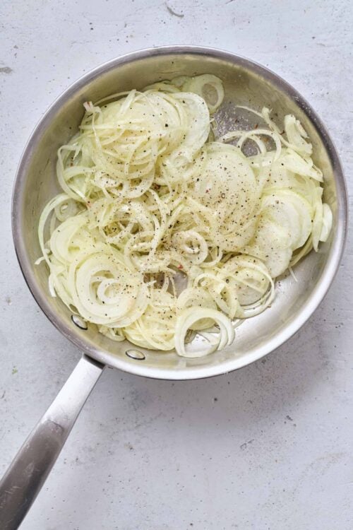 Thinly sliced onions in a stainless steel skillet, seasoned with salt and black pepper.