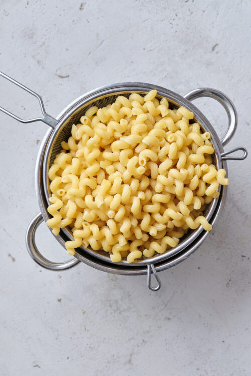 Creamy cooked cavatappi pasta in a stainless steel strainer on a light gray countertop.