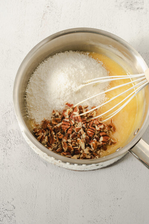Creamy coconut and chopped pecans being mixed into batter with a hand whisk in a stainless steel bowl.