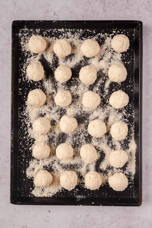 Cream cheese balls on baking sheet with powdered sugar and shredded coconut, ready for baking or chilling.