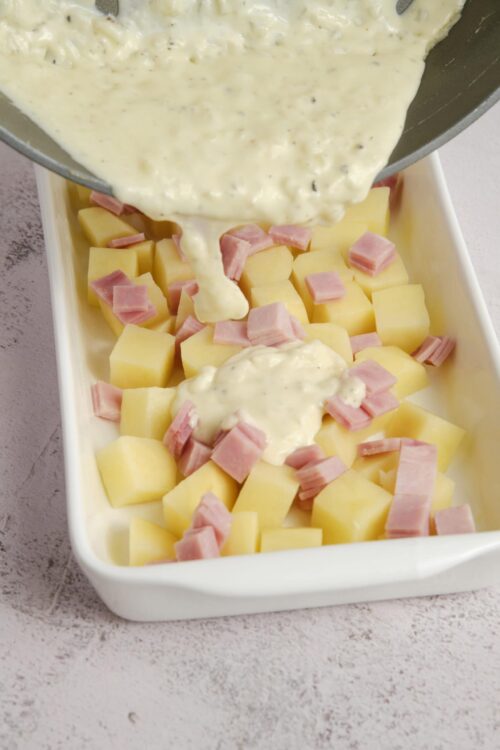 Creamy cheese sauce being poured over diced potatoes and ham in a baking dish.
