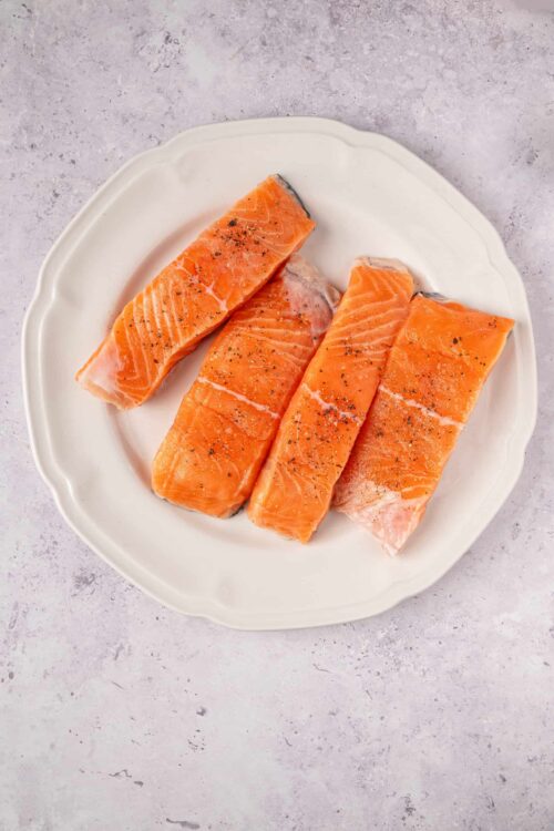 Fresh salmon fillets seasoned with black pepper on a white ceramic plate.