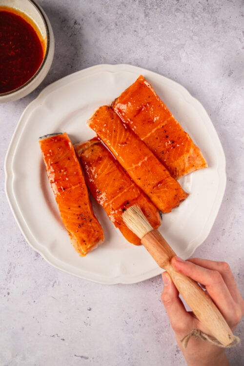 Salmon fillets being brushed with marinade on a white plate with dipping sauce in the background.