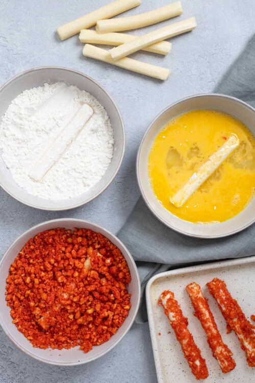 Crispy breaded cheese sticks with flour, egg wash, and seasoned breading ingredients on a gray background.