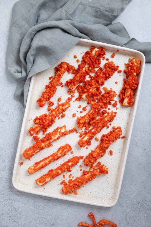 Crispy breaded chicken tenders on a white baking sheet with crumbs and a gray cloth.