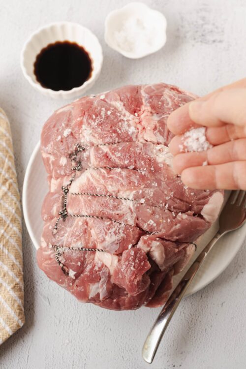 Juicy raw pork shoulder with salt and herbs ready for cooking on a white plate.