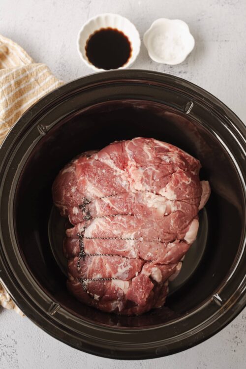 Slow cooker pork roast with seasoning, surrounded by soy sauce, garlic, and salt for flavorful cooking.