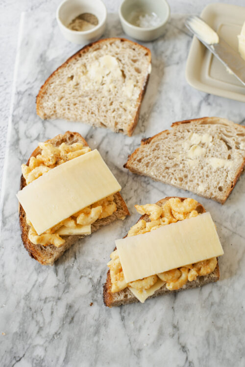 Buttered sourdough bread with macaroni and cheese on top, cheese slices, baking ingredients, marble surface.