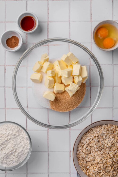 Golden oatmeal cookies ingredients with butter, eggs, oats, cinnamon, vanilla, and dry ingredients on a white tiled kitchen counter.