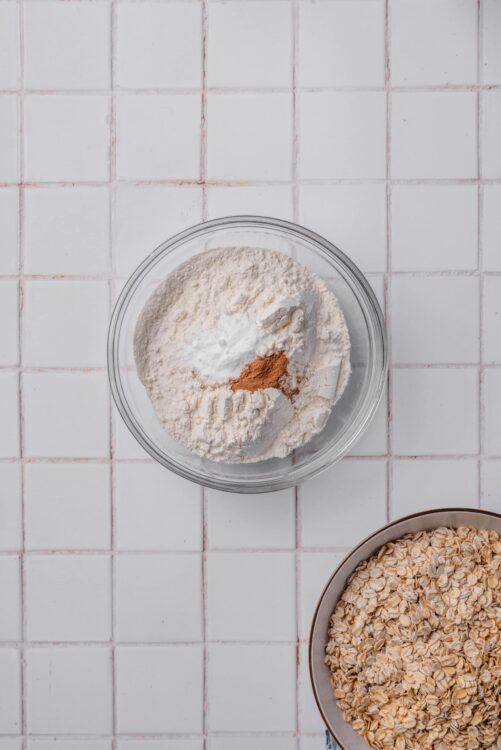 Flour, oats, cinnamon, and baking ingredients on a white tiled kitchen counter.